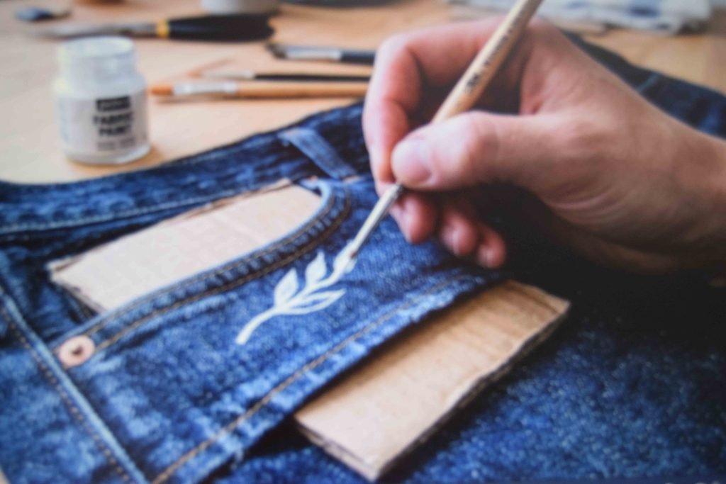 A liner brush applying fabric paint to a denim pocket with cardboard placed inside