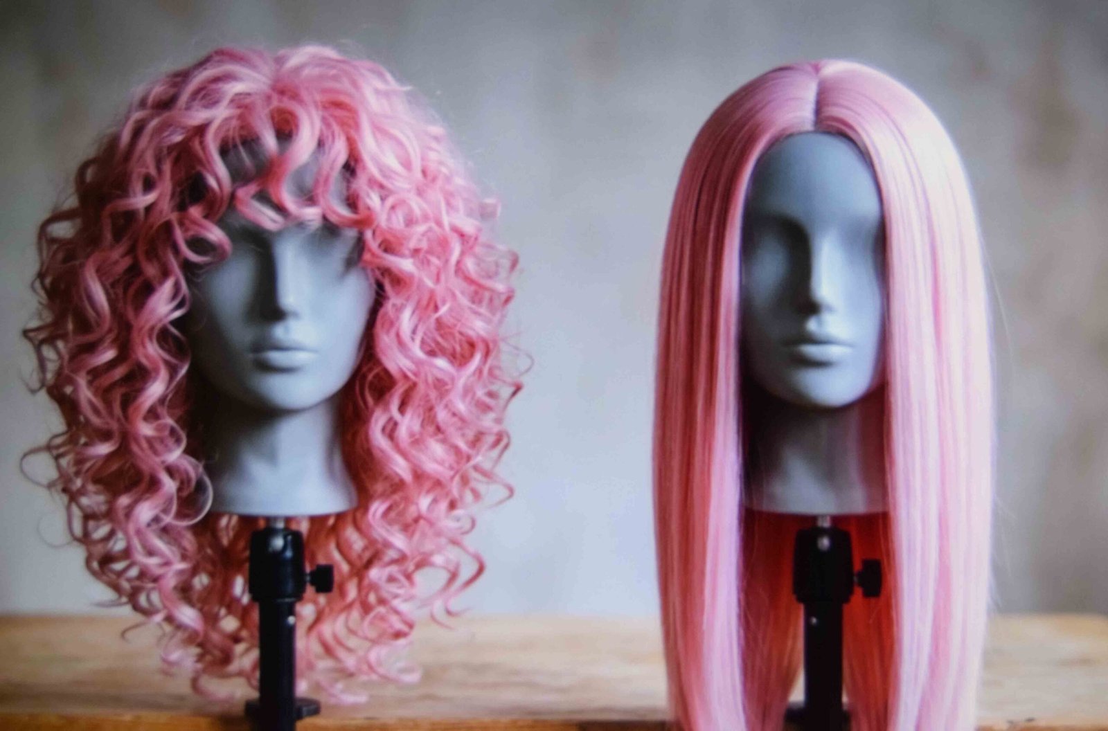 A curly pink wig shown beside a straight one on wig stands