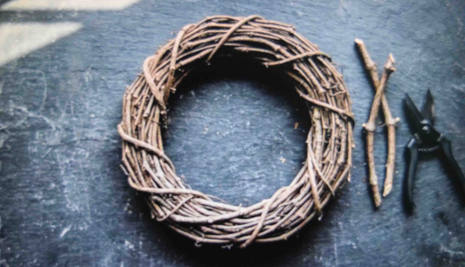 Natural eighteen inch grapevine wreath base lying on a dark work table next to metal floral pruning shears.