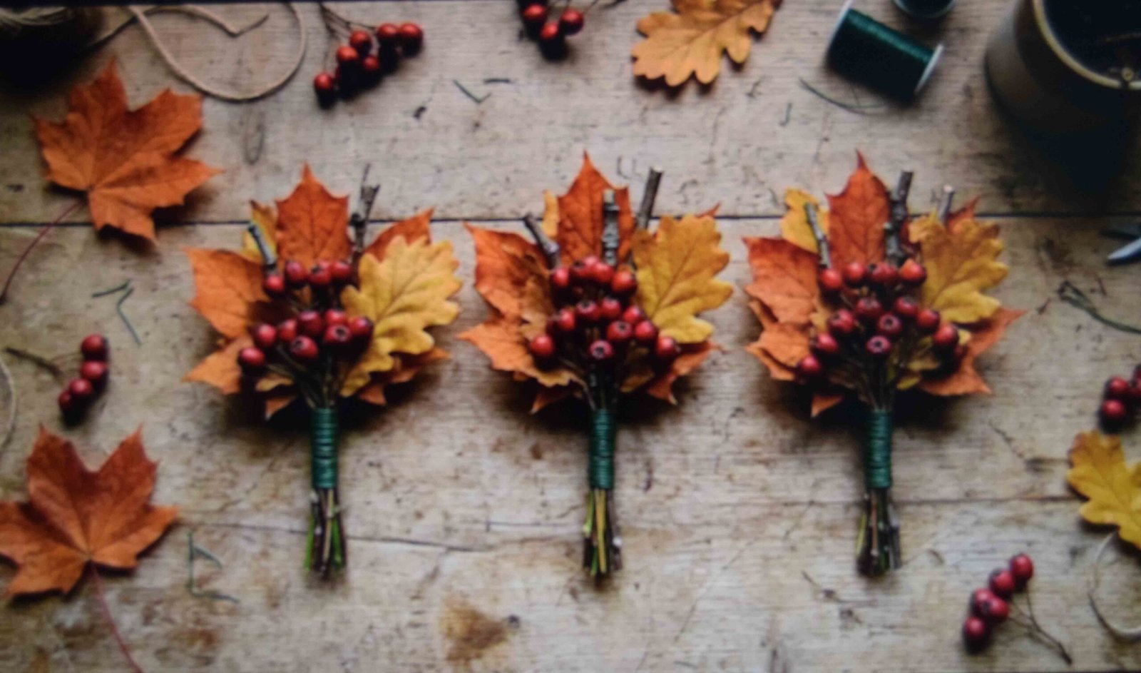 Three pre-assembled faux autumn foliage bundles with maple leaves and berries bound with floral wire on a wood table.