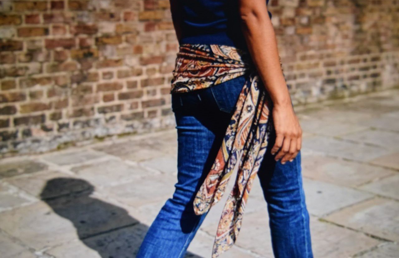 Long printed scarf tied pareo style at the hip over straight leg jeans seen from behind